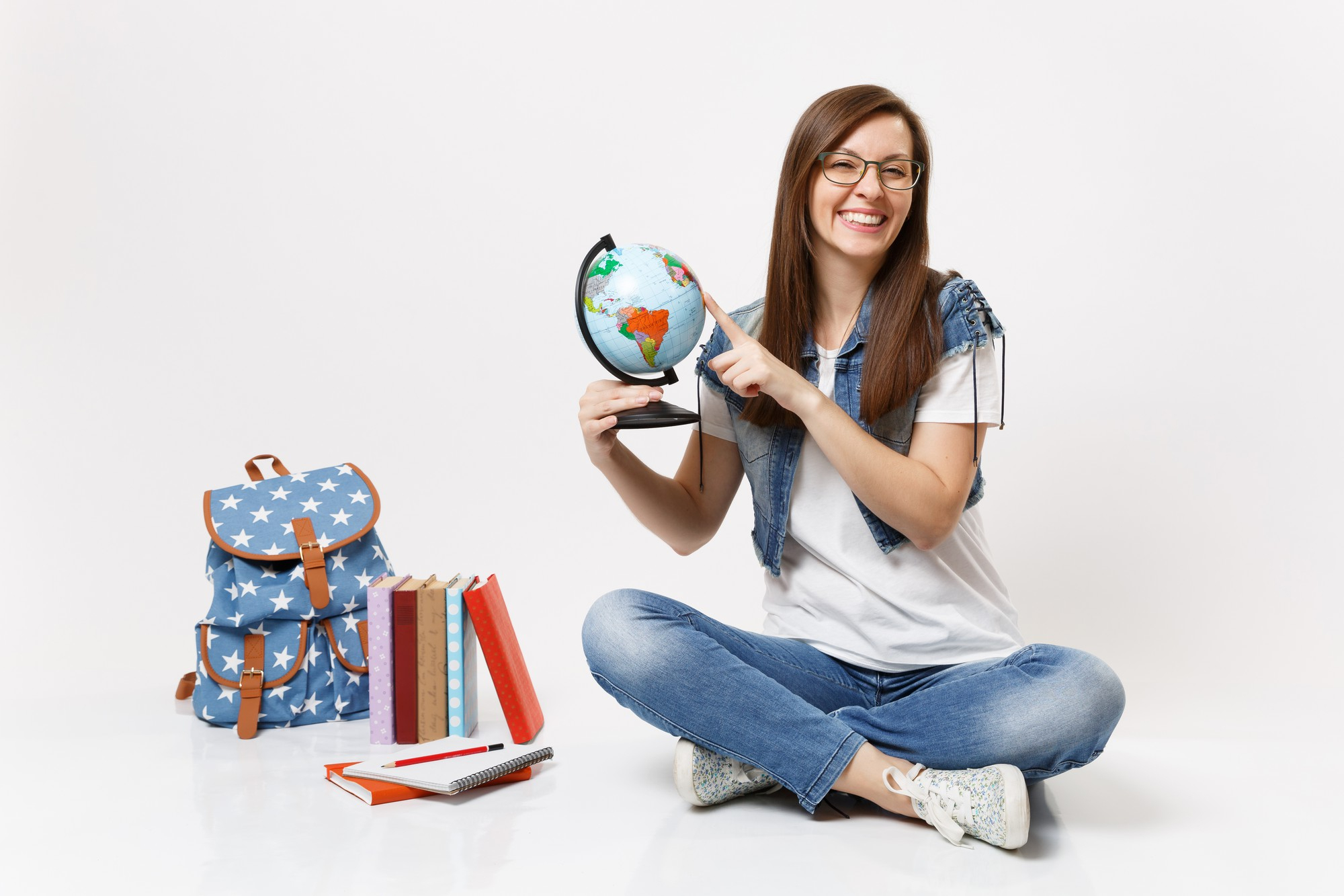 young joyful woman student glasses holding world globe pointing finger countries sitting near backpack school books isolated